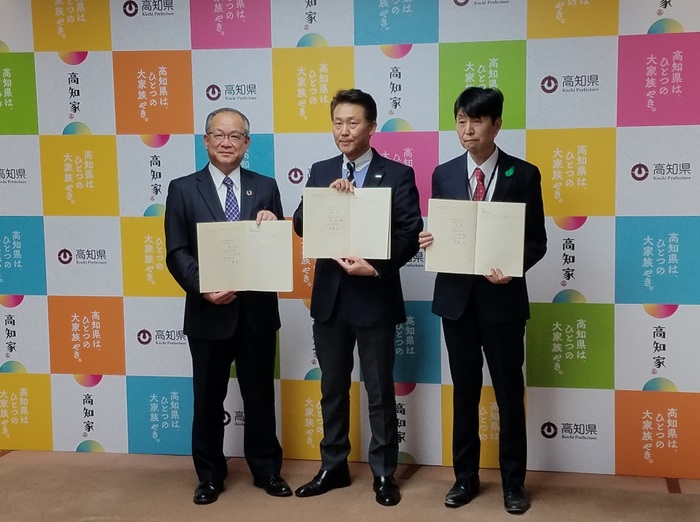 3人の男性が高知県や高知家と記載されているパネルの前でノートを披露している写真