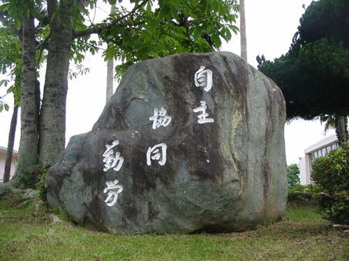 植木が植樹された校庭に、校訓の自主 協同 勤労と書かれた大きな石碑が設置されている写真