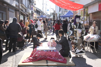 たくさんの人が行き交う商店街の路面に設置されたこたつに入りながら三人親子が食事をしている写真