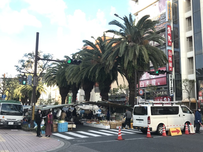 横断歩道のある道路の真ん中に屋根のある出店が並んでいる日曜市の写真