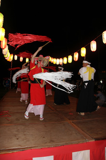 左右に提灯があり赤い浴衣を着ている女性が手結盆踊りをしている様子の写真