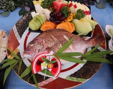 色鮮やかな大皿の真ん中に蒸した丸ごとの鯛、その横に切った果物が添えられ、笹の葉や寿の飾りがあしらわれた料理の写真