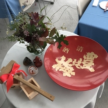 白い丸テーブルの上に、赤い下地に金色の文字で豊能梅と書かれた杯、鏡開き用木づち、飾り花等が置かれている写真