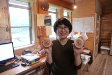 アイビーログ工房内で、眼鏡をかけた女性が高知県産の杉を使ったコースターを両手に持って笑顔で写っている写真。