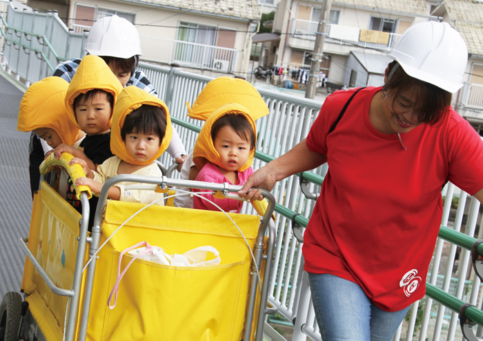 女性と男性の保育士が幼児5人を乗せたカートを引っ張っている写真