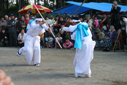 白い袴を着た男性2人が観客たちの前で棒踊りをしている写真
