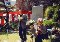 岡本弥太の詩碑の前で一人の男性がマイクの前に立ち歌い、もう一人の男性が椅子に座ってギターを弾いている写真