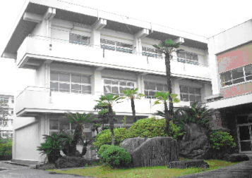 花壇に大きな岩や植木が植えられ、奥に白壁でL字型の3階建ての校舎が建っている建物外観の写真